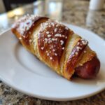 A single, freshly baked pretzel dog wrapped in shiny, golden-brown dough and topped with coarse salt crystals.