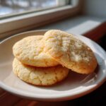Three golden brown sugar cookies stacked slightly on a white plate, illuminated by window light.