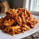 A close-up of a mound of saucy, shredded pulled chicken piled high on a white rectangular plate.