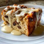 A close-up of a moist slice of bread pudding recipe studded with raisins, topped with thick vanilla sauce.