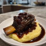 A single, tender braised short ribs topped with a dark, glossy sauce and dried fruit, served over creamy yellow polenta.