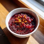 A white bowl filled with rich, dark red homemade cranberry sauce, topped with bright orange zest.