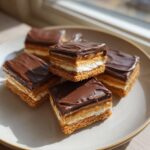 Close-up of layered buckeye graham crackers squares with chocolate, caramel, and marshmallow on a light plate.
