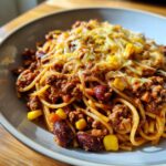 A close-up of a bowl of cheesy enchilada pasta mixed with ground meat, kidney beans, and corn, topped with melted shredded cheese.