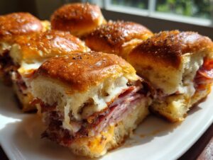 Close-up of several baked grinder sliders filled with melted cheese, ham, and pepperoni on a white plate.