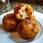 Three golden-brown, deep-fried rice balls stacked on a white plate, one broken open showing melted cheese.