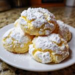 A stack of soft, yellow Christmas gooey butter cookies heavily dusted with white powdered sugar on a white plate.