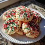 A stack of freshly baked christmas sprinkle cookies covered in red, green, and white nonpareils on a white plate.