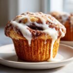 A close-up of a freshly baked cinnamon roll muffin topped with thick white icing dripping down the sides.