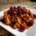 A close-up of shredded cranberry chicken coated in a rich, dark sauce and topped with whole cranberries.