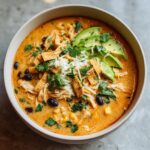 A close-up overhead view of a creamy chicken tortilla soup bowl, garnished with avocado slices, cheese, and tortilla strips.