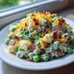 A close-up of creamy pea salad topped with shredded cheddar cheese and crispy bacon bits.