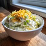 A white bowl filled with creamy pickle dip mixed with celery and topped with shredded cheddar cheese.