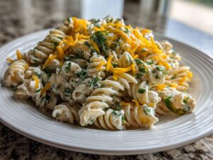 A close-up of creamy pickle pasta salad made with rotini pasta, topped with shredded cheddar cheese and fresh dill.