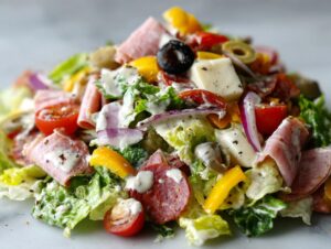 A vibrant close-up of a generous portion of grinder salad featuring lettuce, rolled deli meats, cheese cubes, tomatoes, and creamy dressing.