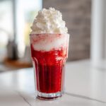 A tall glass of vibrant red strawberry milk topped high with whipped cream.