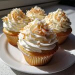 Four freshly baked coconut cupcakes topped with white frosting and toasted coconut flakes, sitting on a white plate.