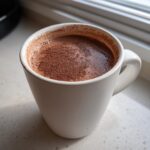 Close-up of a white mug filled with rich, dark hot cocoa mix topped with cocoa powder.
