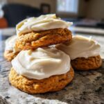 A stack of soft, orange pumpkin cookie halves generously topped with thick white cream cheese frosting.