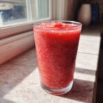 A tall glass filled with a bright red, icy strawberry daiquiri mixture sitting on a sunlit countertop.