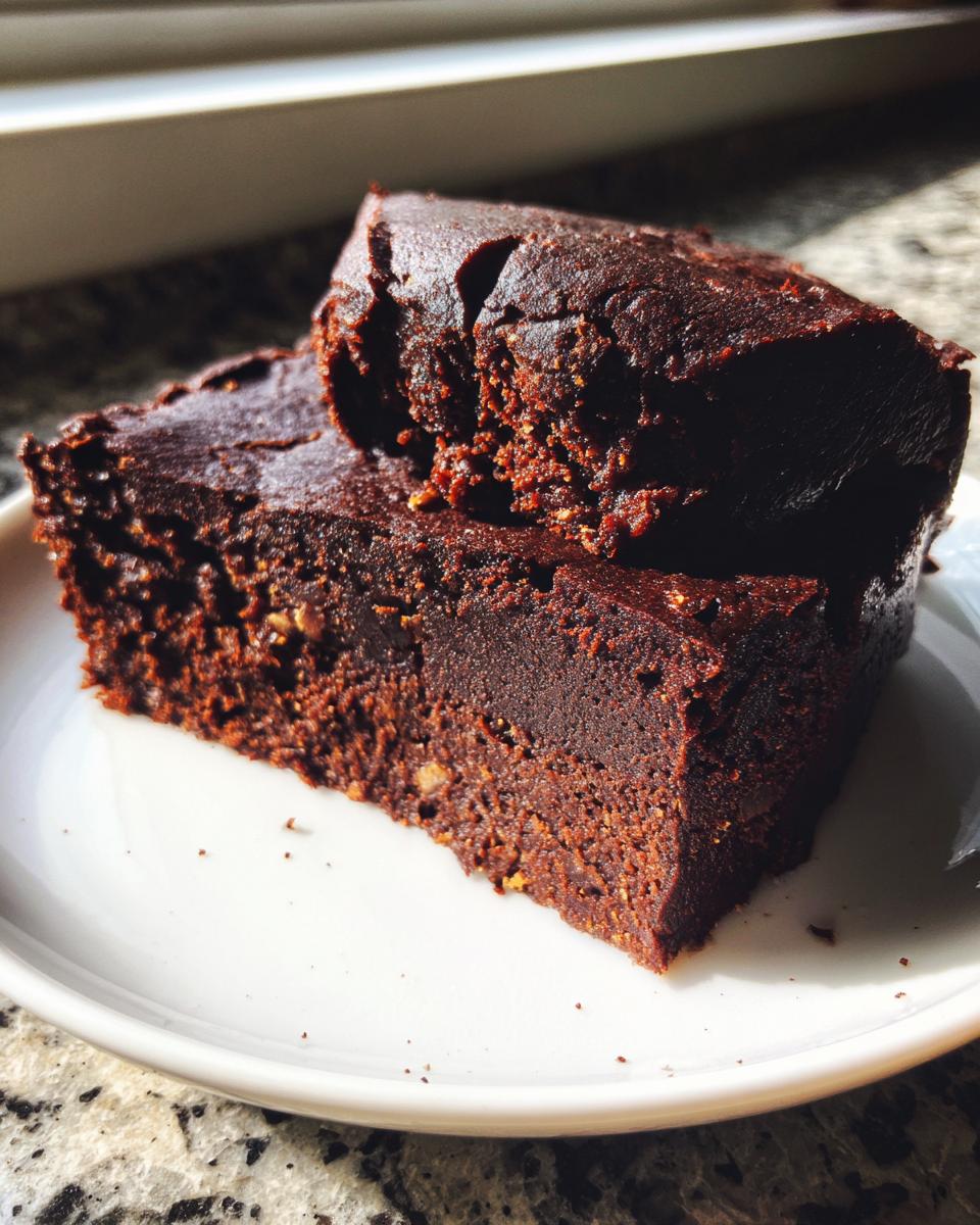 Two rich, dark chocolate date brownies stacked slightly on a white plate, showing a dense, fudgy texture.