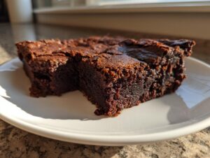 Close-up of a rich, fudgy slice of date brownies on a white plate, highlighted by sunlight.