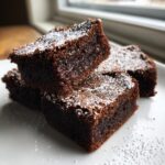 Close-up of three fudgy gingerbread brownies stacked on a white plate, dusted with powdered sugar.
