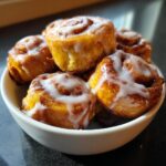 A close-up of several gooey cinnamon roll bites topped with white icing, piled in a small white bowl.