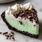 A close-up slice of mint green grasshopper pie with a dark chocolate crust, topped with whipped cream and chocolate shavings.