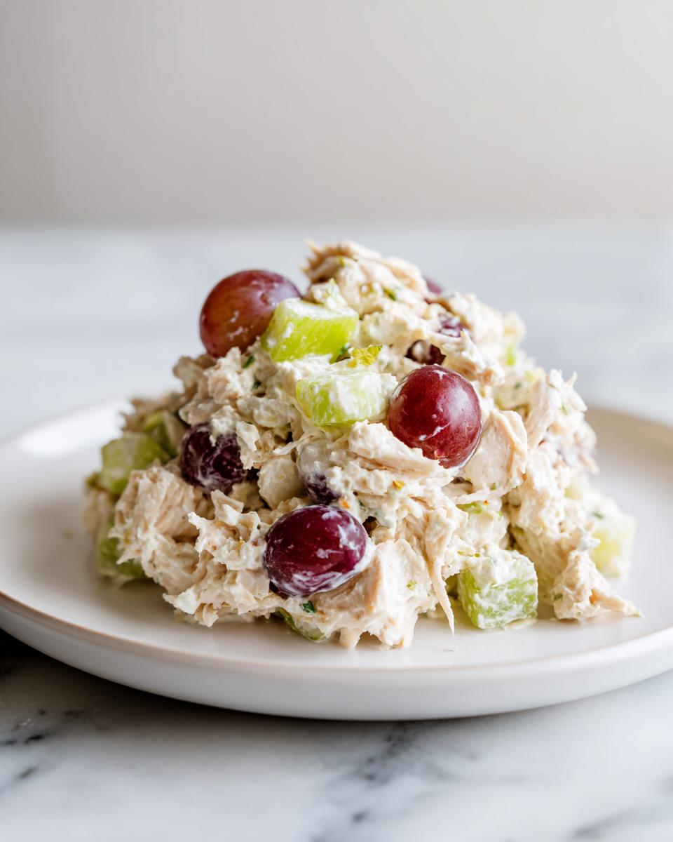 A mound of creamy greek yogurt chicken salad mixed with red grapes and celery on a white plate.