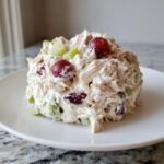 A mound of creamy greek yogurt chicken salad mixed with red grapes and celery on a white plate.