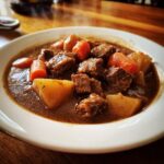 A close-up of a rich, dark brown pork stew served in a white bowl, featuring tender chunks of meat, potatoes, and carrots.