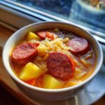 Close-up of a bowl of rich sauerkraut soup featuring thick slices of smoked sausage and chunks of potato.