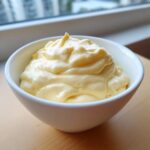 A close-up of thick, creamy, pale yellow homemade mayonnaise served in a small white bowl.