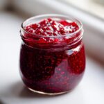 Close-up of a glass jar filled with vibrant, chunky homemade raspberry jam, sitting on a white surface.