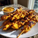 A plate piled high with grilled, golden-yellow chicken satay skewers served next to a small bowl of peanut dipping sauce.