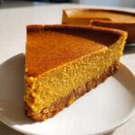 A close-up of a rich, orange slice of keto pumpkin cheesecake with a dark crust on a white plate.