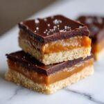 Two stacked, decadent twix bars featuring a shortbread base, thick caramel layer, and dark chocolate top sprinkled with sea salt.