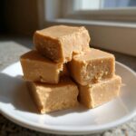 A stack of five creamy, light brown squares of homemade maple fudge resting on a small white plate.