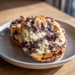 Close-up of two marbled chocolate chip cheesecake cookies with a crumbly top texture on a light plate.