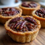 A close-up photo of several freshly baked mini pecan pies with glistening filling and whole pecans on top, resting on a wooden surface.