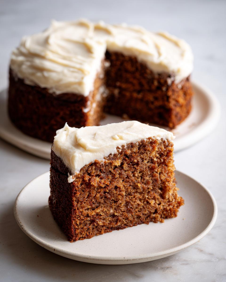 A close-up slice of moist applesauce cake topped with thick cream cheese frosting, with the rest of the cake blurred in the background.
