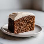 Close-up of a moist, dark slice of applesauce cake topped with thick white frosting on a small plate.