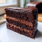 Close-up of a moist, dark slice of chocolate fudge cake with two layers of filling and glossy chocolate ganache frosting.