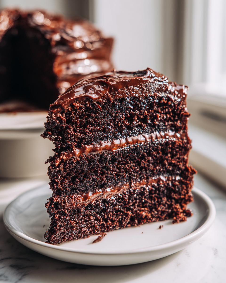 A thick, moist slice of dark chocolate fudge cake with rich frosting layers served on a white plate.