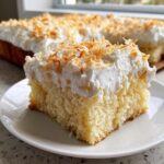 A close-up of a moist slice of coconut poke cake topped with whipped cream and toasted coconut flakes.
