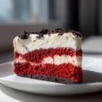 A rich slice of red velvet cheesecake featuring layers of bright red cake and white cream cheese filling, topped with dark cookie crumbs.