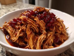 Close-up of shredded cranberry chicken coated in sauce and topped with whole cranberries in a white bowl.
