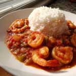 A close-up of a serving of rich shrimp creole served alongside a mound of white rice on a white plate.