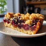 A close-up of a rich slice of plum cake with a thick, juicy fruit layer and a buttery crumble topping.